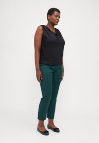 Černý top bez rukávů s výstřihem do cowl, zkombinovaný s tmavě zelenými slim-fit kalhotami. Model má na sobě černé baleríny. Hladká textilie.