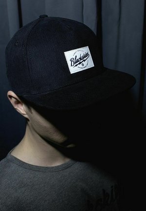 Person trägt eine schwarze Blackskies-Kappe und ein graues Hemd, das Gesicht größtenteils schattiert, dunkler Hintergrund.