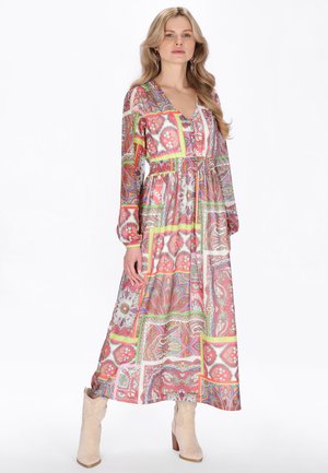 Donna che indossa un abito lungo a maniche lunghe con fantasie colorate, scollo a V e stivali beige alla caviglia, in piedi davanti a uno sfondo semplice.