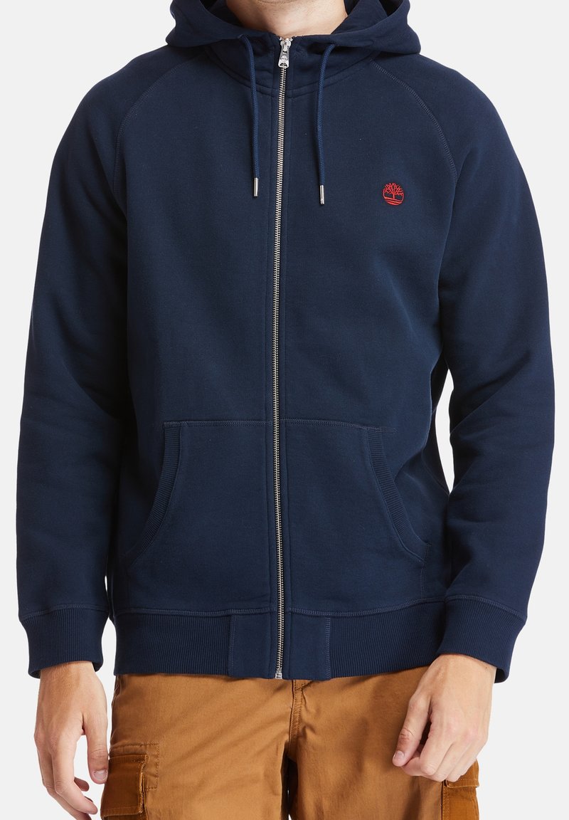 Homme portant un sweat à capuche bleu marine zippé avec poches avant et un petit logo d'arbre rouge sur la poitrine, associé à un pantalon marron clair.