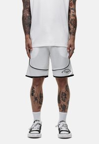 Pantaloni corti bianchi in rete con accenti neri, caratterizzati da un orlo curvo e il logo del marchio. Indossati con calze bianche e scarpe nere, che mostrano tatuaggi dettagliati.