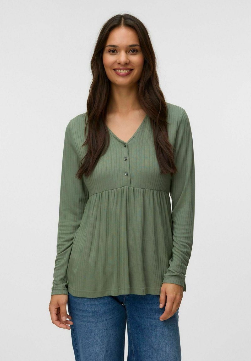 Jeune femme aux longs cheveux foncés portant un haut vert côtelé à manches longues avec des boutons et un jean bleu, souriant à la caméra.