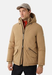Beige isolierte Jacke mit Kapuze, mit einem Reißverschluss auf der Vorderseite, zwei schrägen Taschen und akzentuierenden Details. Getragen über einem roten Pullover und dunklen Jeans.