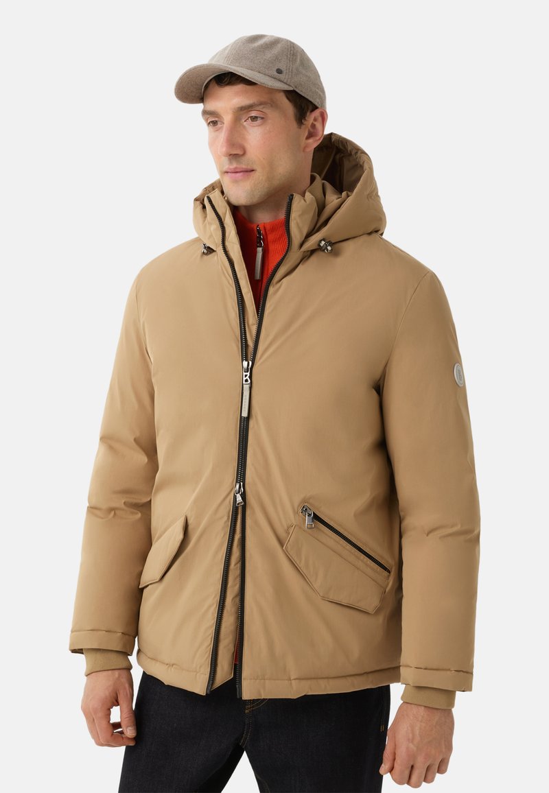 Beige isolierte Jacke mit Kapuze, mit einem Reißverschluss auf der Vorderseite, zwei schrägen Taschen und akzentuierenden Details. Getragen über einem roten Pullover und dunklen Jeans.