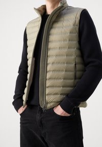 Gilet imbottito verde oliva con trapuntatura orizzontale, colletto alto, chiusura con cerniera e tasche laterali, indossato sopra un maglione nero e jeans scuri.