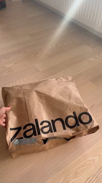 Käsi, mis hoiab kokku tõmmatud pruunist paberist paketti, millel on mustade tähtedega kirjas "zalando", asetatud heledale puidust põrandale radiaatori lähedale.