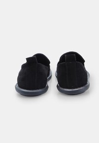 O pereche de pantofi negri din suede, tip slip-on, văzuți din spate, care arată cusăturile și banderolele de prindere pe un fundal alb.