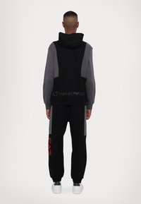 Svart och grå sweatshirt med huva och logotyp på baksidan, kombinerad med lösa svarta byxor med en röd grafisk detalj på sidan.