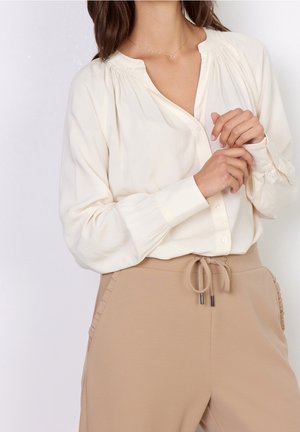 Femme portant un chemisier blanc à manches longues et un pantalon beige à cordons, les mains délicatement jointes devant, sur un fond uni.