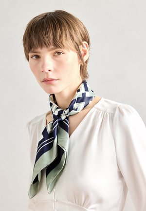 Ung person med kort brunt hår iført en hvid bluse og et navyblåt og grønt tørklæde med geometrisk mønster bundet om halsen.