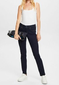 Žena v tmavě modrých skinny džínách, bílém tílku a bílých teniskách, držící černou psaníčko proti jednobarevnému pozadí.