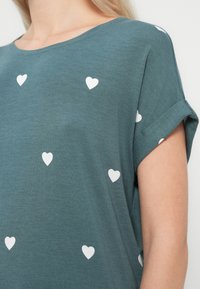 Τυρκουάζ πουκάμισο με κοντά μανίκια και μικρά λευκά σχέδια καρδιάς, με διπλωμένη μανσέτα και μερική προβολή του λαιμού και του μπράτσου του φορέα.