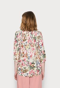 Kvinna med axellångt hår som bär en vit blommig blus med 3/4 ärmar och rosa byxor, vänd bort mot en enfärgad ljus bakgrund.