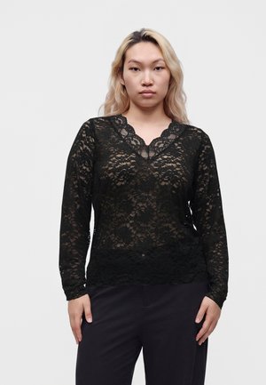 Frau mit langen blonden Haaren, die ein schwarzes Spitzen-Longsleeve und eine schwarze Hose trägt, steht vor einem schlichten hellen Hintergrund.