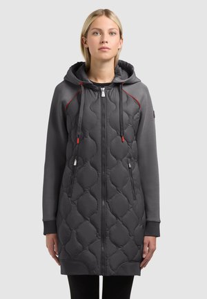 Młoda kobieta zwrócona przodem, ubrana w długą, ciemnoszarą, pikowaną kurtkę z kapturem, z czerwonymi sznureczkami i zasuwanymi kieszeniami.