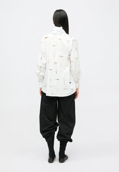 Γυναίκα με μακριά καστανά μαλλιά, ντυμένη με λευκό πουκάμισο μακρυμάνικο με ακανόνιστα κοψίματα, μαύρο άνετο παντελόνι και μαύρα παπούτσια, στέκεται γυρισμένη πλάτη.