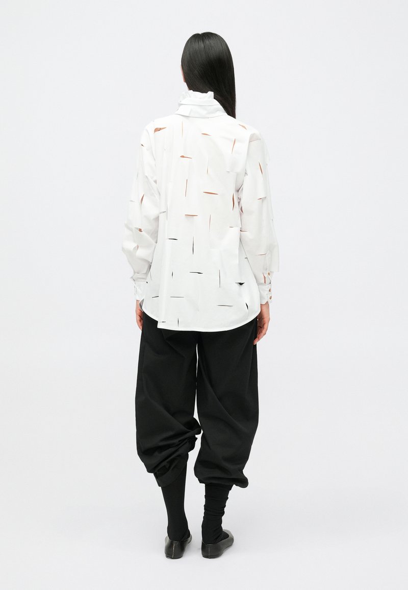 Kvinna med långt mörkt hår som bär en vit långärmad tröja med oregelbundna utskärningar, svarta lösa byxor och svarta skor, stående med ryggen mot kameran.