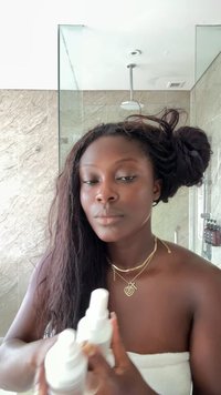 Due bottiglie spray bianche sono tenute in mano. La persona ha capelli lunghi, mossi e scuri, indossa un asciugamano bianco e collane d'oro a strati.