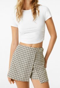 Weißes kurzärmliges Cropped-Top kombiniert mit einem Rock im schwarz-weißen Hahnentrittmuster, der über eine seitliche Öffnung und eine kreisförmige Schnallendetail verfügt.