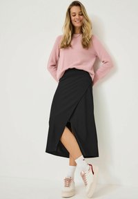 Jupe noire à enroulement avec une fente sur le côté, associée à un pull en laine léger rose pâle et des baskets blanches avec des accents roses. Matériau lisse, look décontracté.