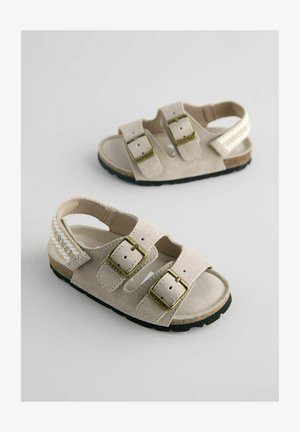 Beige mokasiner av ruskinn med to justerbare stropper, gullfargede spenner og en teksturert såle. Utstyrt med flettet detaljering og logo.