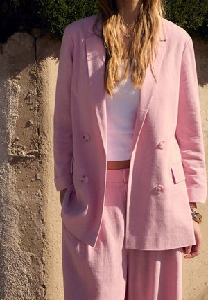 Frau trägt ein hellrosa zweireihiges Blazer und passende Hose über einem weißen Crop-Top, steht vor einer strukturierten beigen Wand.