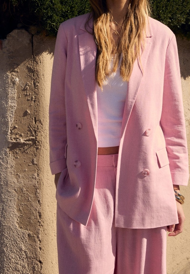 Nainen, jolla on päällä vaaleanpunainen tuplanepparikauluksellinen bleiseri ja siihen sopivat housut valkoisen crop topin päällä, seisoo teksturoidun vaalean beigen seinän edessä.