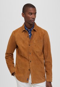 Camisa de ante marrón con botón en el frente, cuello puntiagudo, bolsillo en el pecho y un corte relajado, llevada sobre una camisa de cuello azul.