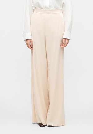 Kobieta w beżowych spodniach z wysokim stanem i szerokimi nogawkami, w białej, długorękawowej bluzce oraz czarnych butach, stojąca na tle jednolitego tła.