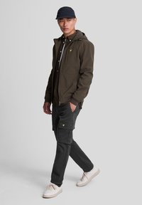 Olive green jacket with hood and front pockets, worn over a brown shirt. Black cargo pants feature side pockets, complemented by white sneakers.