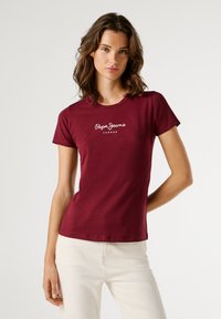 Mladá žena s vlnitými hnedými vlasmi, oblečená v priliehavom burgundskom tričku Pepe Jeans a svetlých béžových nohaviciach, stojí na jednofarebnom pozadí.