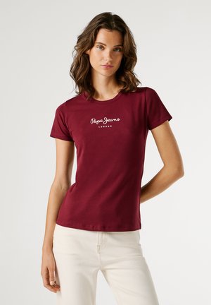 Jonge vrouw met golvend bruin haar, gekleed in een getailleerde bordeauxrode Pepe Jeans t-shirt en lichtbeige broek, staand tegen een effen achtergrond.