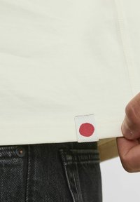 Cremfarbendes T-Shirt mit kurzen Ärmeln, das ein strukturiertes Etikett mit einem roten Punkt aufweist. Über dunklen Jeans getragen, die Nahtdetails zeigen.