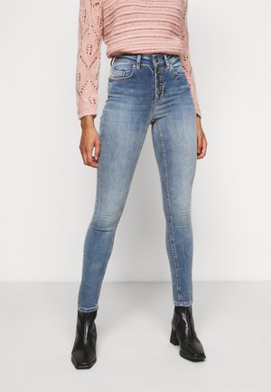 Femme portant un jean skinny bleu clair taille haute avec des boutons à l'avant, un pull en maille rose et des bottines noires, debout sur un fond blanc.
