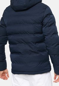 Granatowa kurtka puchowa z poziomym przeszyciem, kapturem i elastycznymi mankietami. Posiada gładką fakturę oraz lekko błyszczące wykończenie.
