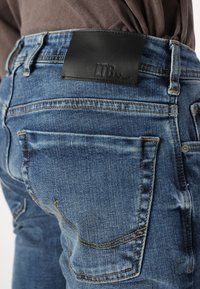 Jeans ajustados de color azul hechos de denim con una etiqueta de cuero negro en la cintura. Presenta bolsillos traseros con costuras visibles. Acabado ligeramente desgastado.