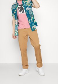 Persoon in een teal patroon kortemouwshirt over een roze graphic T-shirt, beige broek en witte sneakers, staand tegen een witte achtergrond.