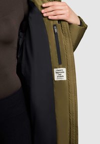 Hand som håller upp en olivgrön jacka som avslöjar inre foder i svart, en dragkedjeficka och en tygetikett som säger "En varm kram gjord av tyg."