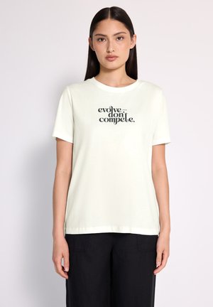 Femme aux longs cheveux bruns portant un t-shirt blanc avec le texte "évoluez, ne vous battez pas" et un pantalon noir, se tenant devant un fond clair.
