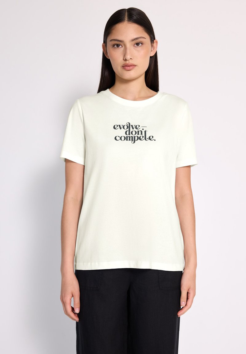Femme aux longs cheveux bruns portant un t-shirt blanc avec le texte "évoluez, ne vous battez pas" et un pantalon noir, se tenant devant un fond clair.