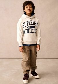 Bílá mikina s kapucí a potiskem "SUPERDRY ATHLETICS", béžové cargo kalhoty a maroon vysoké tenisky. Texturovaná tkanina a uvolněný střih.