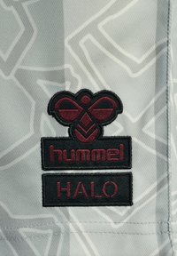 Textilmärket har en mörk vinröd logotyp med en stiliserad design ovanför ordet "hummel" och "HALO" i fet stil, mot en ljusgrå mönstrad bakgrund.