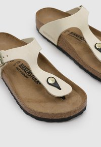 Infradito Birkenstock color crema con suola in sughero, cinturino regolabile e un dettaglio di toe post nero con accentuazione metallica.