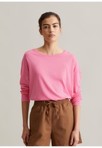 Frau mit zurückgebundenem dunklem Haar, die ein langärmliges pinkes Shirt und braune Hosen mit Kordelzug trägt, steht vor einem schlichten Hintergrund.