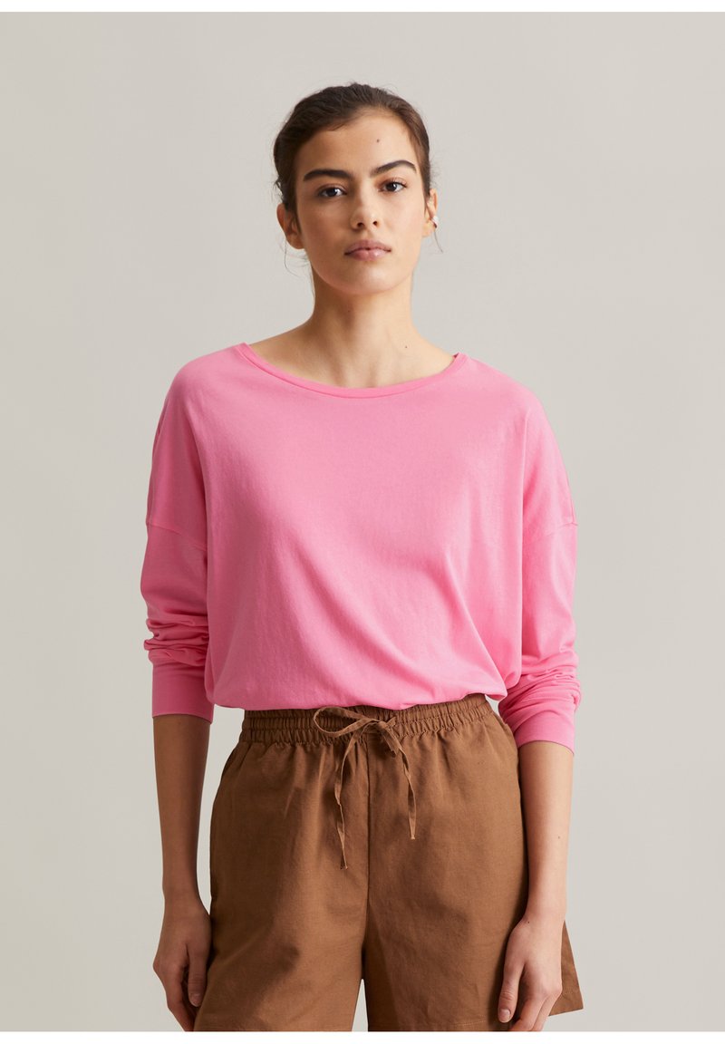 Frau mit zurückgebundenem dunklem Haar, die ein langärmliges pinkes Shirt und braune Hosen mit Kordelzug trägt, steht vor einem schlichten Hintergrund.