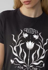 Zwarte katoenen t-shirt met een witte bloemgrafiek met het woord "GRATEFUL" erboven en "FOR THE" eronder, gebogen tekst en vloeiende plantontwerpen.