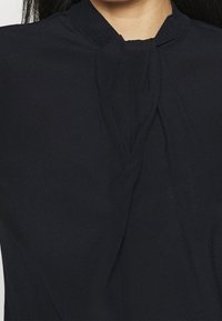 Blouse noire avec un col montant agrémenté d'un détail noué à l'avant, confectionnée en tissu lisse à la texture douce.