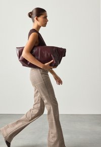 Borsa oversize in pelle bordeaux con texture liscia, abbinata a top marrone aderente e pantaloni a zampa a quadri, in un ambiente minimalista.