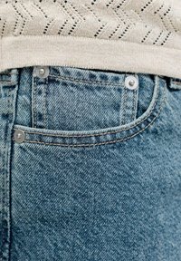 Béžový pletený sveter zastrčený do modrých džínsov, zobrazuje predné vrecko s kovovými nitmi a detailmi švov.