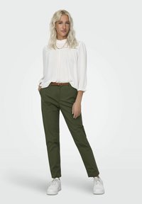 Blusa bianca a maniche lunghe con scollatura arricciata, abbinata a pantaloni olive verdi a sigaretta e sneakers bianche, con una cintura intrecciata marrone.